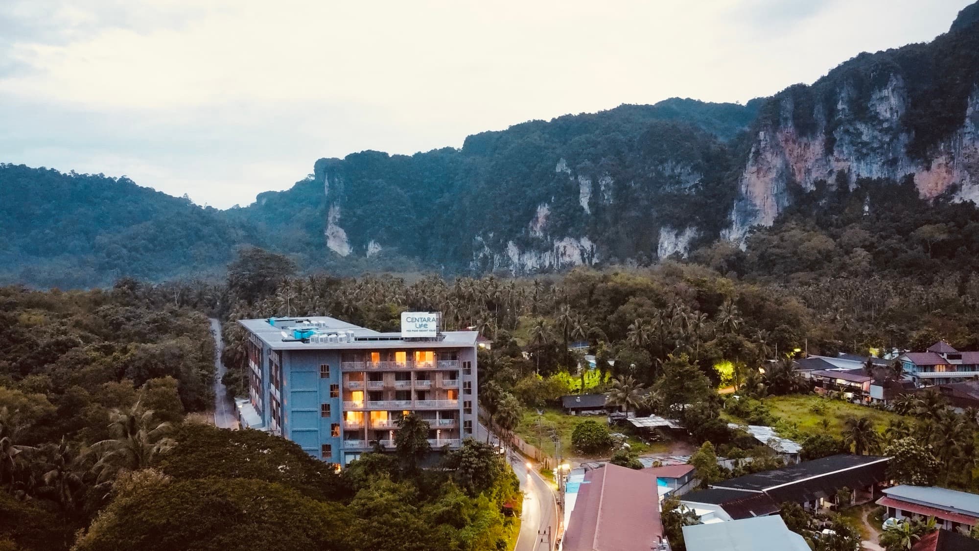 A hotel beneath forested limestone cliffs photographed from the air at dusk