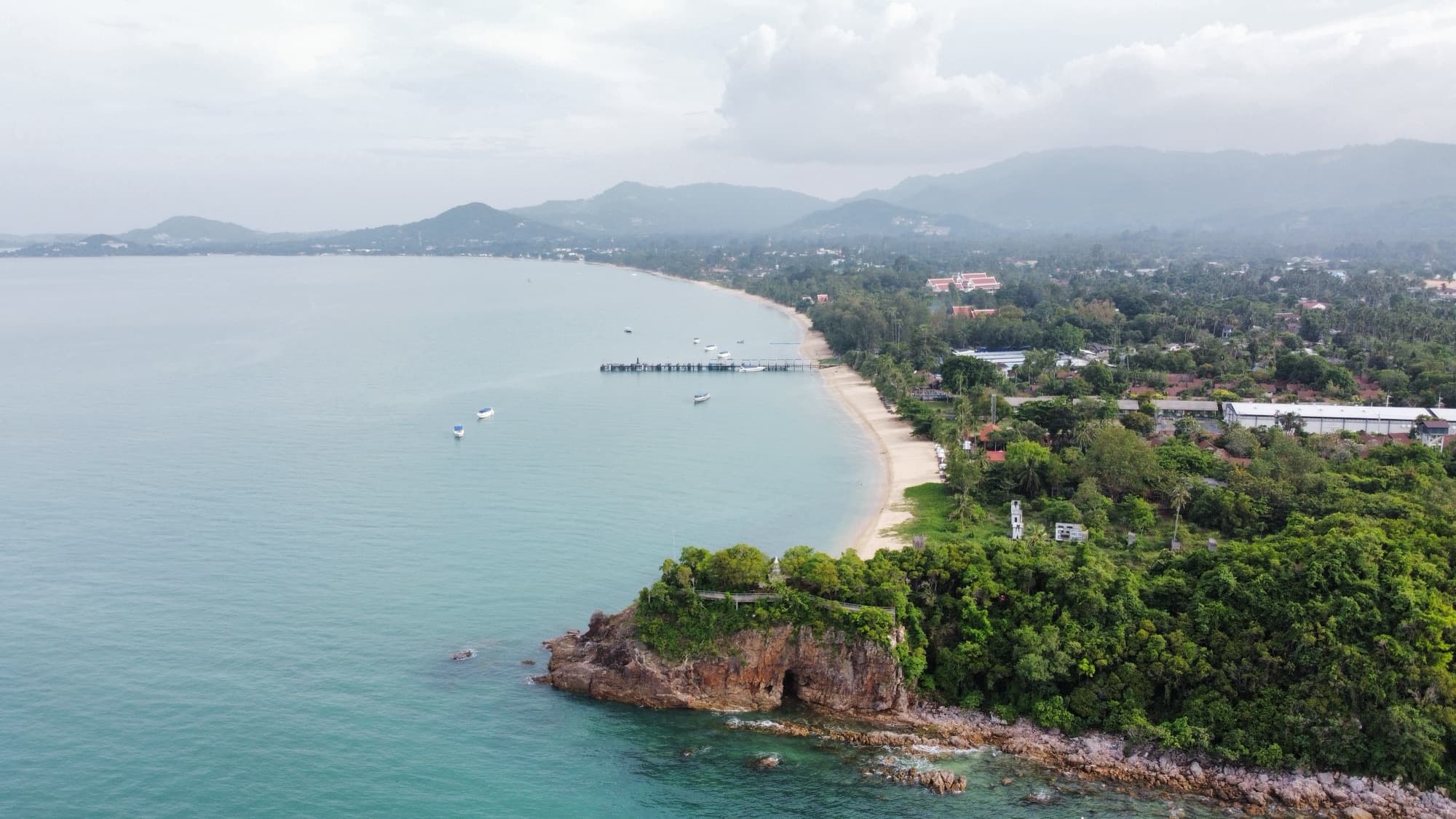 A curving coastline, pier and moored boats photographed from the air